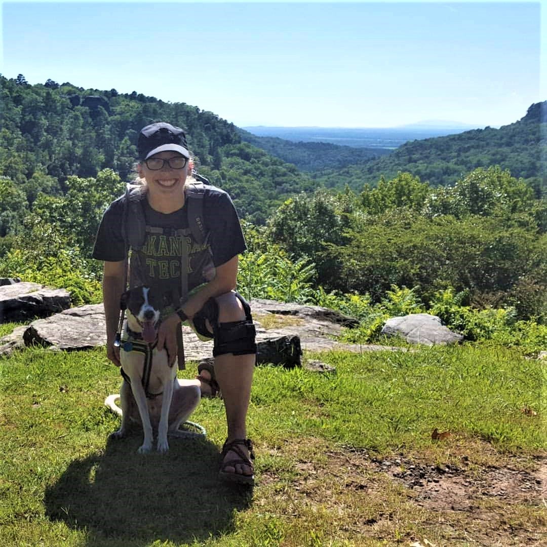 A hiker kneels in the grass with her sitting dog.